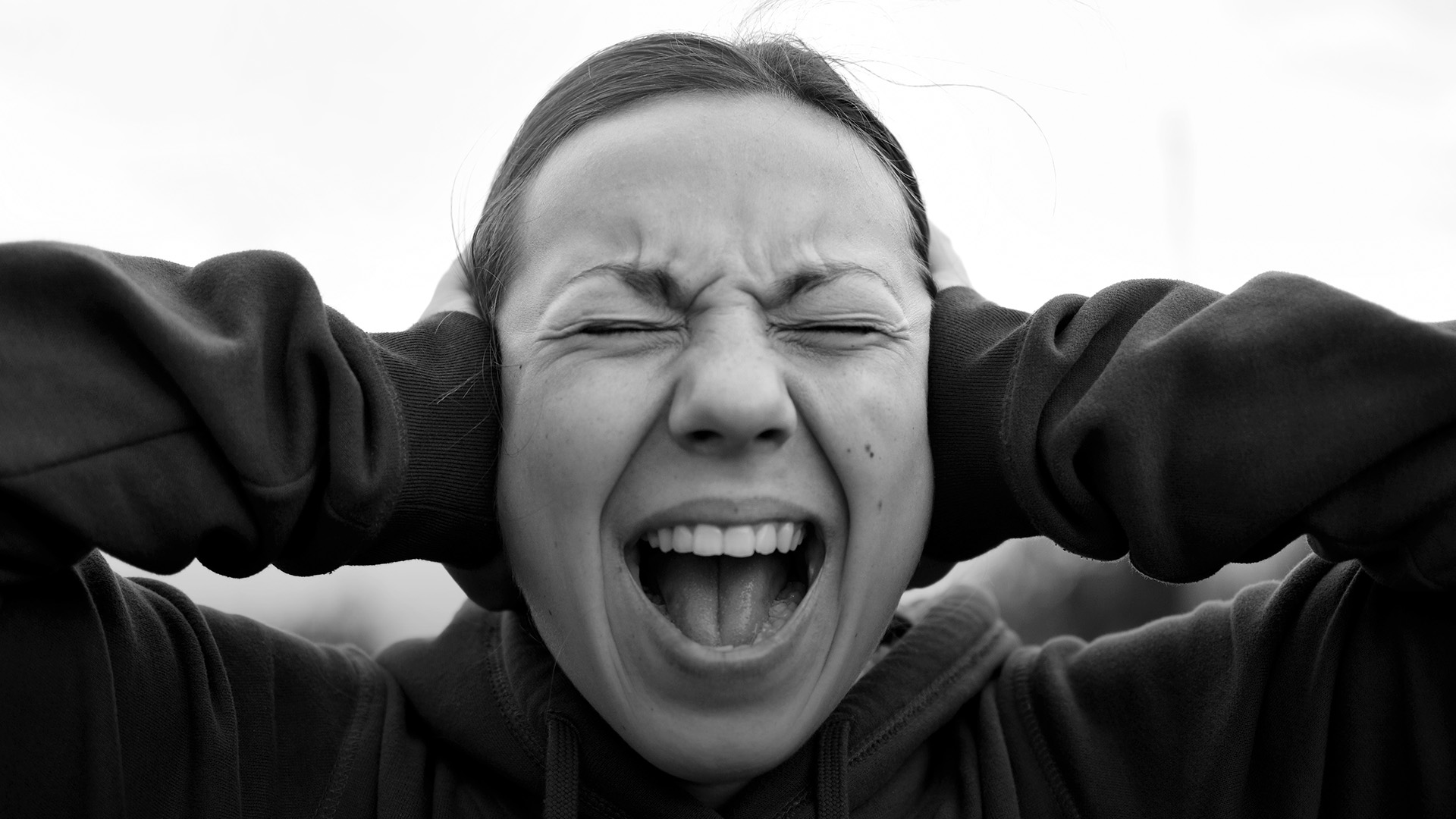 A young woman screams with her mouth open while covering her ears with her hands.
