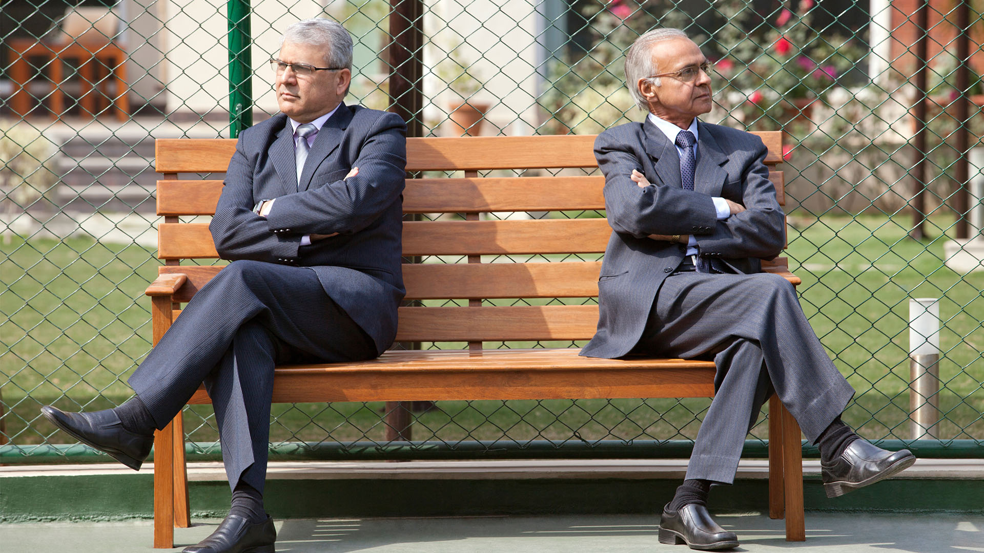 Two businessmen sit on bench, crossing their arms